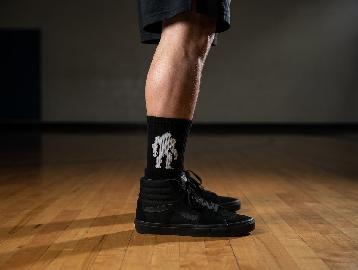 Massquatch black crew sock with white walking sasquatch silhouette, worn with black Vans SK8-Hi — moody gym hardwood hero.