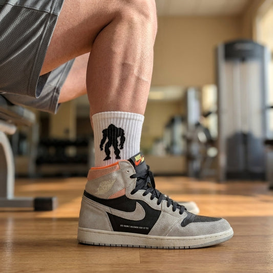 Massquatch white crew sock with black walking sasquatch silhouette, worn with Air Jordan 1 Retro High OG Neutral Grey — cinematic practice gym hero.